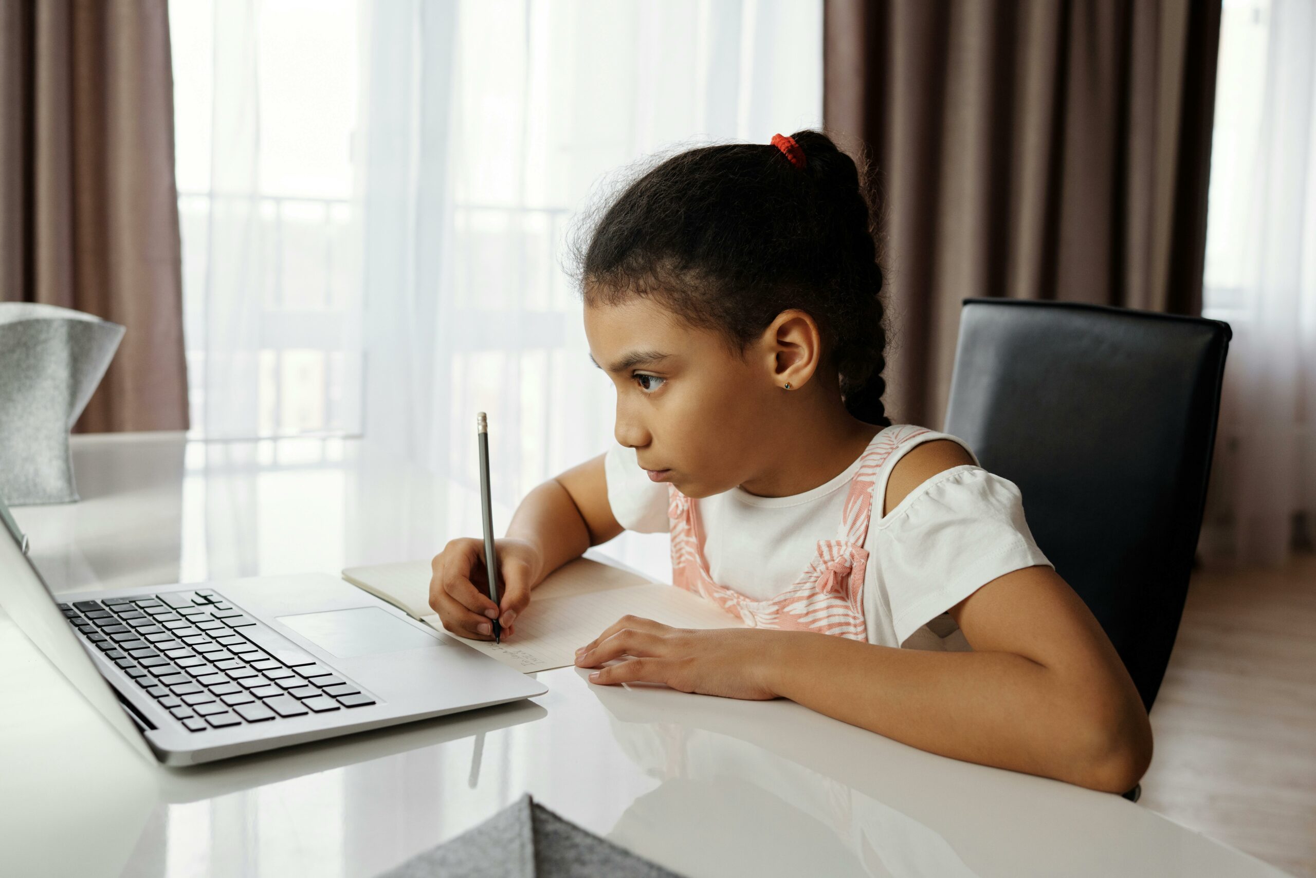 child-sitting-at-a-desk-in-front-of-laptop-writing-on-a-notebook